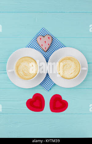 Zwei Tassen Cappuccino, Ansicht von oben. Zusammensetzung mit ein paar Kaffeetassen, herzförmige Cookie und rotem Papier Herzen. Valentines Tag Holz- Hintergrund. Stockfoto