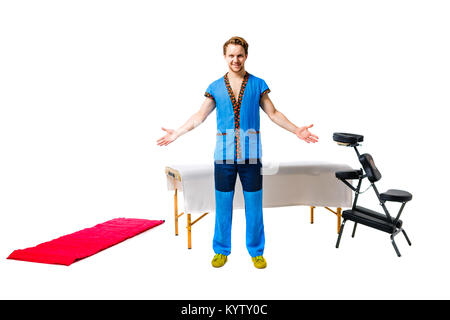 Männlichen masseur in der blauen Uniform steht mit der Geste des Grußes Hände zu den Seiten mit Smile neben Anlagen für Massageliege, Klappstuhl, rote Wolldecke o Stockfoto