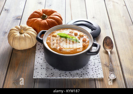 Leckere Kürbissuppe in Schwarz Keramik Topf auf rustikalen, mit Holz. Die Suppe mit Creme, Croutons und Basilikumblätter serviert. Stockfoto