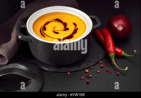 Pikante Kürbis creme Suppe serviert in Keramik Topf mit Kürbiskernöl. Vegetarisches Gericht für den Herbst Zeit gewürzt mit Chili und roten Zwiebeln. Stockfoto