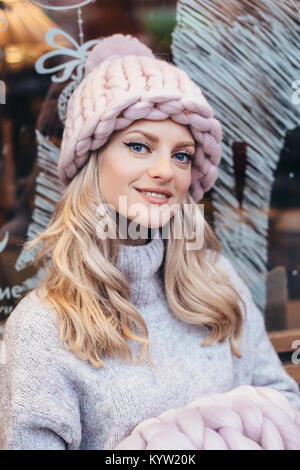 Junge blonde Frau in Rauchigen rosa Strickmütze machen posiert auf einer Holzbank mit Weihnachten Cafe - Fenster im Hintergrund Stockfoto