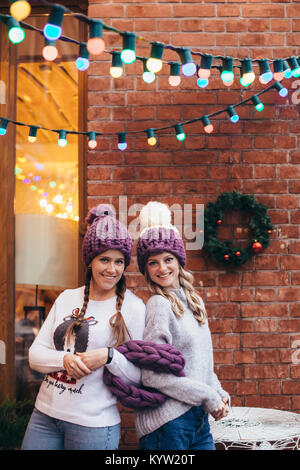 Zwei Frauen in Lila gestrickte Hüte, Lila infinity Schal und blaue Jeans mit Mauer und Weihnachten Girlande auf dem Hintergrund posiert Stockfoto