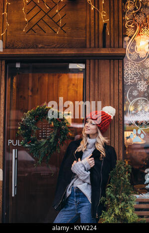 Junge blonde Frau in Rot Strickmütze mit pompon, grau Pullover, schwarze Jacke und Jeans machen posiert mit Christmac Fenster Cafe auf dem hinterg Stockfoto