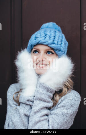 Junge blonde Frau in Blau Strickmütze, weiße und flauschige Fausthandschuhe und graue Strickjacke mit Holztür auf dem Hintergrund posiert Stockfoto