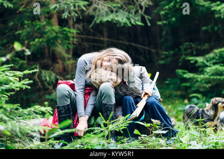 Hugging Kinder sitzen in der Waldlichtung, den Kopf geneigt. Bruder und Schwester spielen im Clearing von Bäumen und mit Hund umgeben. Famil Stockfoto