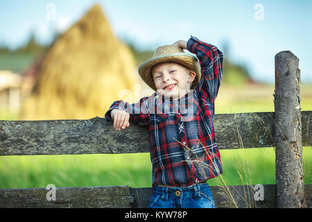 Close up Portrait von kleinen Jungen in Hut mit Stroh in Mund auf hölzernen Zaun lehnte. Happy chilhood Konzept. Ferienhäuser Stockfoto