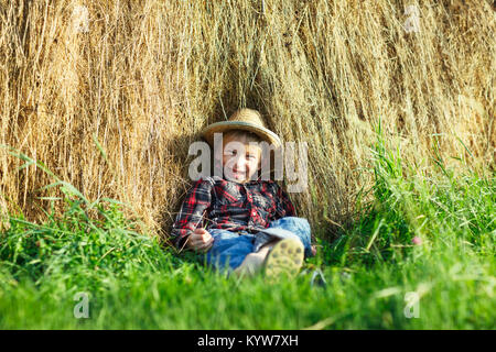 Happy Boy im Stroh Hut in Heuhaufen sitzen, Draußen, schaut die Kamera. Lächelnd kind Heuhaufen auf grünem Gras unter der heißen Sonne entspannen. Junger Mann verbringt Ho Stockfoto