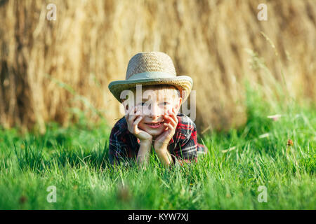 Schöner kleiner Junge liegt auf Gras auf dem Hintergrund von haystack. Kind liegt in der Natur, liegt auf dem Bauch und propps Kopf mit Händen. Porträt der jungen Stockfoto