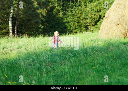 Junge in hat läuft Gras zu Wald, Ansicht von hinten. Junge im roten Hemd und Jeans durch die Wiese geht an Dickicht, und auf der rechten Seite ist heuschober. Fröhliche vacatio Stockfoto