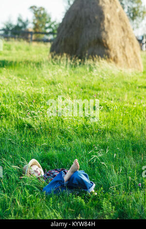 Schöner kleiner Junge liegend im Gras auf dem Rücken, die Hände hinter den Kopf. Kleine Träumer in der Wiese mit Hut liegt, die Augen geschlossen. Kind Schlafen und lächelnd in der Natu Stockfoto