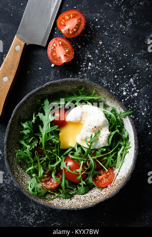 Gesunder Salat mit Ruccola, Tomaten und pochiertem Ei in der Schüssel auf dunklem Hintergrund. Ansicht von oben mit Platz für Text kopieren Stockfoto