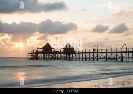 Hölzerne Seebrücke und Strohdächern an einem tropischen Strand bei Sonnenaufgang, Insel Sansibar Stockfoto