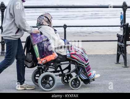 Jüngere Person (Pflege/Familie?) Rollstuhl schieben mit älteren Frau entlang der Küste. Großbritannien Stockfoto