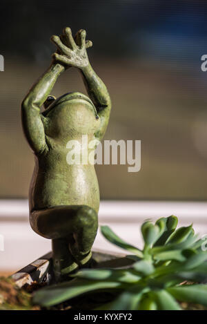 Yoga Frosch in die Baumhaltung. Figur Statue mit saftigen Haus im sonnigen Fenster. niedlich Grün kleine Objekt mit den Händen in der Luft lächelnd. Soft Focus. Stockfoto