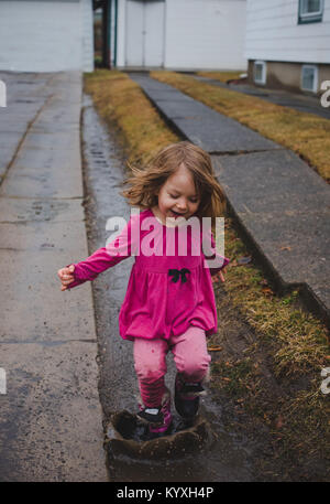 Kleinkind Mädchen mit rosa, Springen in ein Schlammloch in einer Fahrstraße an einem regnerischen Tag. Stockfoto