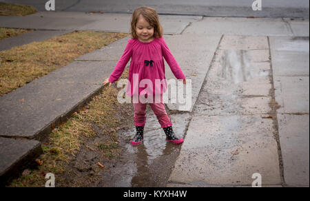 Ein Kleinkind trägt ein rosafarbenes Hemd, rosa Hose und Rosa regen Stiefel steht in einem schlammloch in einer Fahrstraße an einem regnerischen Tag. Stockfoto