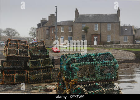 Stapel von Hummer Töpfe oder Reusen auf Stein Pier, die Häuser der St. Margaret's Hope, Orkney, und ein rotes Ruderboot im Hintergrund auf einem grauen regnerischen Tag. Stockfoto