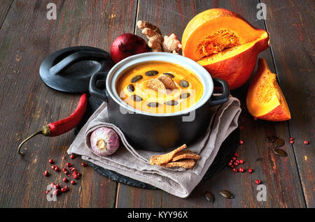 Pikante Kürbis creme Suppe mit Ingwer, spanische Zwiebeln, Knoblauch und Chili in keramischen Pfanne auf dunklem Holz. Die Suppe mit Croutons, Creme und pumpki serviert. Stockfoto