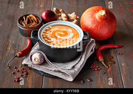 Pikante Kürbis creme Suppe mit Ingwer, spanische Zwiebeln, Knoblauch und Chili in keramischen Pfanne auf dunklem Holz. Die Suppe mit Croutons und Sahne serviert. Stockfoto