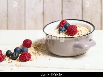 Haferflocken in grau Topf und frische Himbeeren und Blaubeeren auf weissem Holztisch Stockfoto