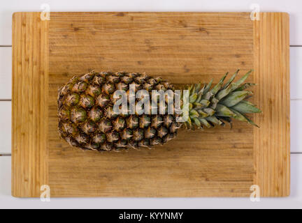 Ganze reife Ananas mit Blättern, die auf einer hölzernen Schneidebrett über eine rustikale weiß Holz Tisch gesehen von oben bis schließen Stockfoto