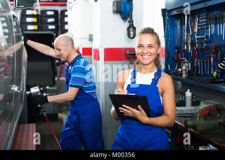 Freundlich lächelnden jungen Frau in Blau insgesamt stehen und schreiben Sie die Daten in auto Werkstatt Stockfoto