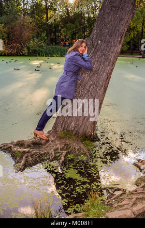 Junge Mädchen steht lehnte sich gegen eine große Bücken Baum im Wasser im Herbst. Stockfoto