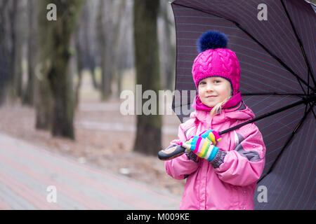 Portrait von wenig liebenswert kaukasische Mädchen mit rosa warme Jacke und Strickmütze wandern unter dem Dach in einem Stadtpark an kalter Tag. Stockfoto