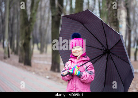 Wenig liebenswert kaukasische Mädchen mit rosa warme Jacke und Strickmütze wandern unter dem Dach in einem Stadtpark an kalter Tag. Stockfoto