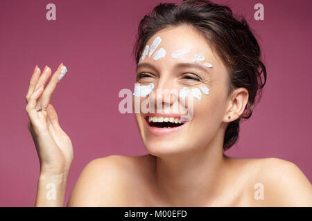 Lachende Mädchen mit makelloser Haut Anwendung Feuchtigkeits-creme auf ihrem Gesicht auf rosa Hintergrund. Gesichtsbehandlung Stockfoto