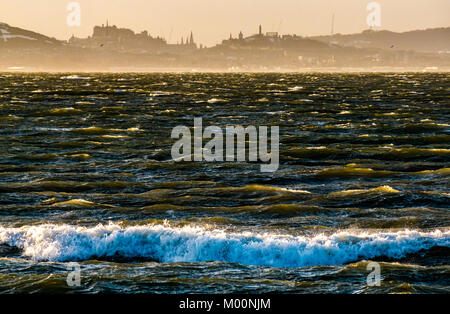 Longniddry Bents, East Lothian, Schottland, Großbritannien. Starke Winde erzeugen Wellen im Firth of Forth. Farbenfroher orangefarbener Sonnenuntergang über dem Firth of Forth mit Blick nach Westen vom Strand in Longniddry auf den markanten Grundriss der Skyline von Edinburgh mit Schloss und Kirchtürmen in Edinburgh Stockfoto