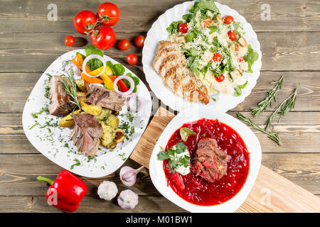 Verschiedene Gerichte auf Holztisch, Ansicht von oben. Gemüse Rüben Suppe, Lammrücken mit gebratenen Kartoffeln, gegrilltem Hühnchen Caesar Salat. Stockfoto