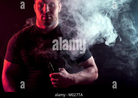 Eine junge elegante Mann Raucher in einem schwarzen T-Shirt ist eine vape in seine Hand und um und eine grosse Rauchwolke aus vape auf einem dunklen isoliert Hinterg Stockfoto