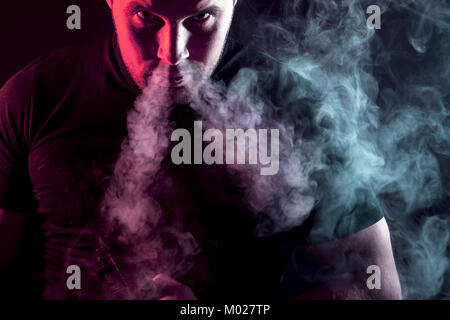 Eine junge stilvolle männliche Raucher in einem schwarzen T-Shirt starrt und atmet aus der Nase eine große Wolke von Rauch aus der vape auf einem dunklen Hintergrund isoliert Stockfoto