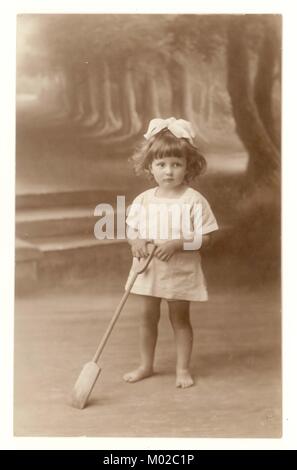 Original Anfang des 20. Jahrhunderts Studio-Porträt-Postkarte mit ländlichen Szene Hintergrund, von niedlichen jungen Mädchen mit einem Band in den Haaren, hält einen Spaten, Urlaubsfoto, ca. Anfang der 1920er Jahre, Southampton, Hampshire, England, Großbritannien Stockfoto