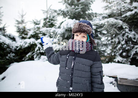 Glückliches Kind in warme Kleidung wirft einen Schneeball an der Kamera. Junge Spaß im Schnee. Stockfoto