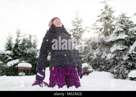Lachende Frau in warme Winterkleidung sitzen auf verschneiten Boden in ihrem Gesicht durch Schnee schlagen. Frau genießen Schnee. Stockfoto