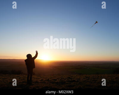 Kind flying a Kite in einem klaren blauen Himmel über der Britischen Landschaft gegen die untergehende Sonne über dem Horizont silhouettiert Stockfoto