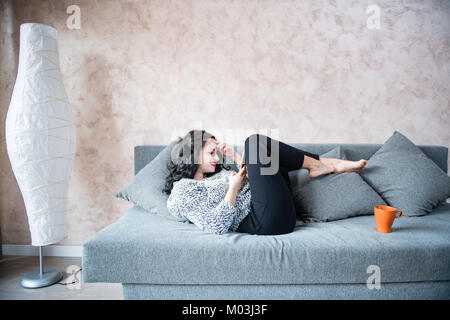 Junge Erwachsene brünette Frau lächelnd auf Smart Phone liegen auf der Couch zu Hause Stockfoto