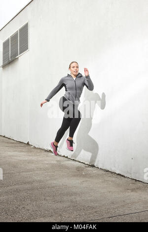 Eine junge Frau, die in der Mitte springen - Luft, draußen, weiße Wand hinter, Einfach, minimalistisch, sportliche Kleidung. Stockfoto