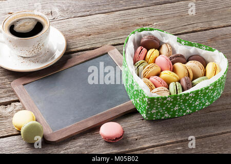 Bunte Makronen in Valentines Tag herzförmigen Geschenkbox und Kaffee Tasse auf hölzernen Tisch. Mit Tafel für Ihre Grußansagen Stockfoto