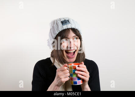 Portrait von glücklichen Frau in wollmütze Lachen (Mund weit geöffnet), mit Becher, wärmende Hände. Aufgeregt, cheesy Grin zeigt Zähne. Stockfoto