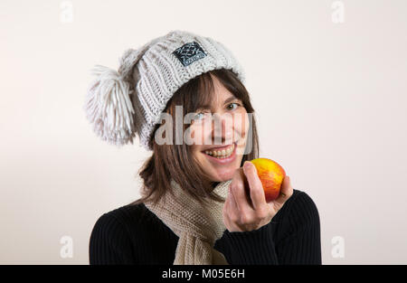 Indoor-Porträt, glücklich gesund isoliert Mädchen lächelnd, in wollig Bommel Hut, hält frischen Apfel in der Hand bereit, einen Bissen zu nehmen. Schönes großes natürliches Lächeln. Stockfoto