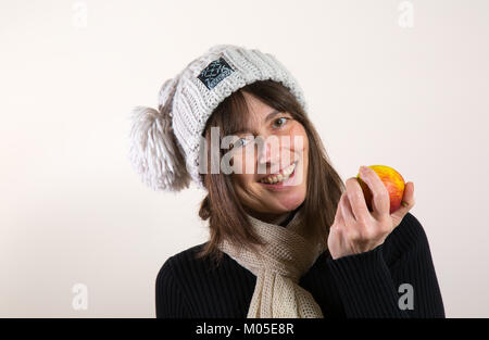 Indoor Portrait von glücklichen, gesunden Langhaarigen weiblichen, in Woolly bobble Hut & Schal, frischen Apfel in der Hand zu nehmen. Schönes, großes natürliches Lächeln. Stockfoto