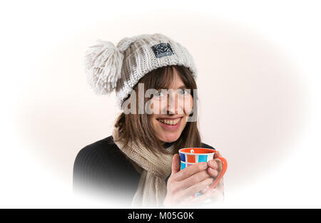 Innenportrait einer britischen Frau, die lächelnd in Wollmütze und Schal auf die Kamera schaut, Kaffee hält und warme kalte Hände auf einem heißen Becher hält. Ein Lächeln auf den Lippen. Stockfoto