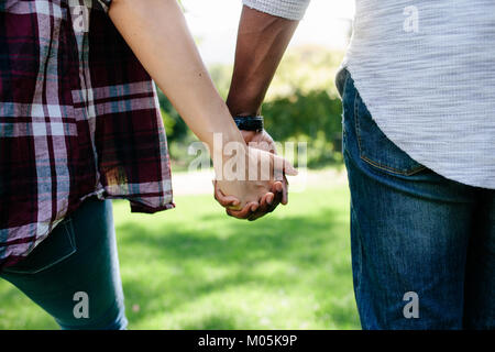 In der Nähe von Paar halten sich an den Händen und gehen im Freien. Rückansicht des Mannes und der Frau halten sich an den Händen der jeweils anderen beim Gehen in einen Park. Stockfoto