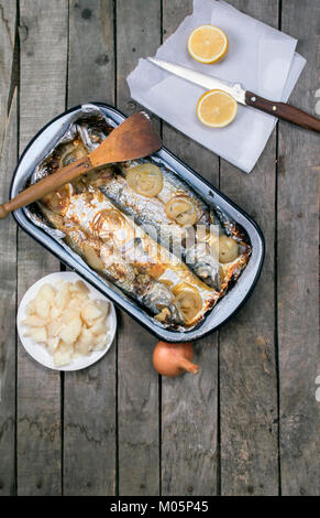 Blick von oben auf die gebackene Makrele fisch Auflauf mit Holz- Schaufel auf alten Holztisch mit ganzen Zwiebel, Zitrone und Kartoffelsalat auf grau Holz- Hintergrund Stockfoto