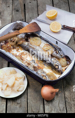 Gebackene Makrele fisch Auflauf mit Holz- Schaufel auf alten Holztisch mit ganzen Zwiebel, geschnitten Zitrone und Kartoffelsalat auf grau Holz- Hintergrund. Stockfoto