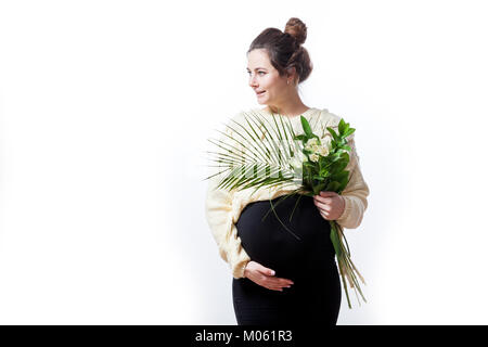 Junge fröhliche dunkelhaarige Frau in Form gestrickter Pullover und schwarzes Kleid auf der späten Schwangerschaft hält einen Strauß Blumen und lächelnd auf Weiß isoliert backgro Stockfoto