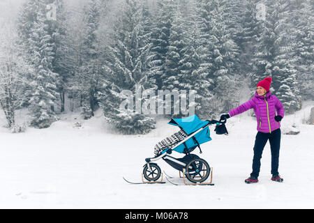 Mutter mit Baby Stroller genießen Mutterschaft im Winter Wald, in den Bergen. Joggen oder Walken Frau mit Schlitten pram in Wäldern. Das Stockfoto